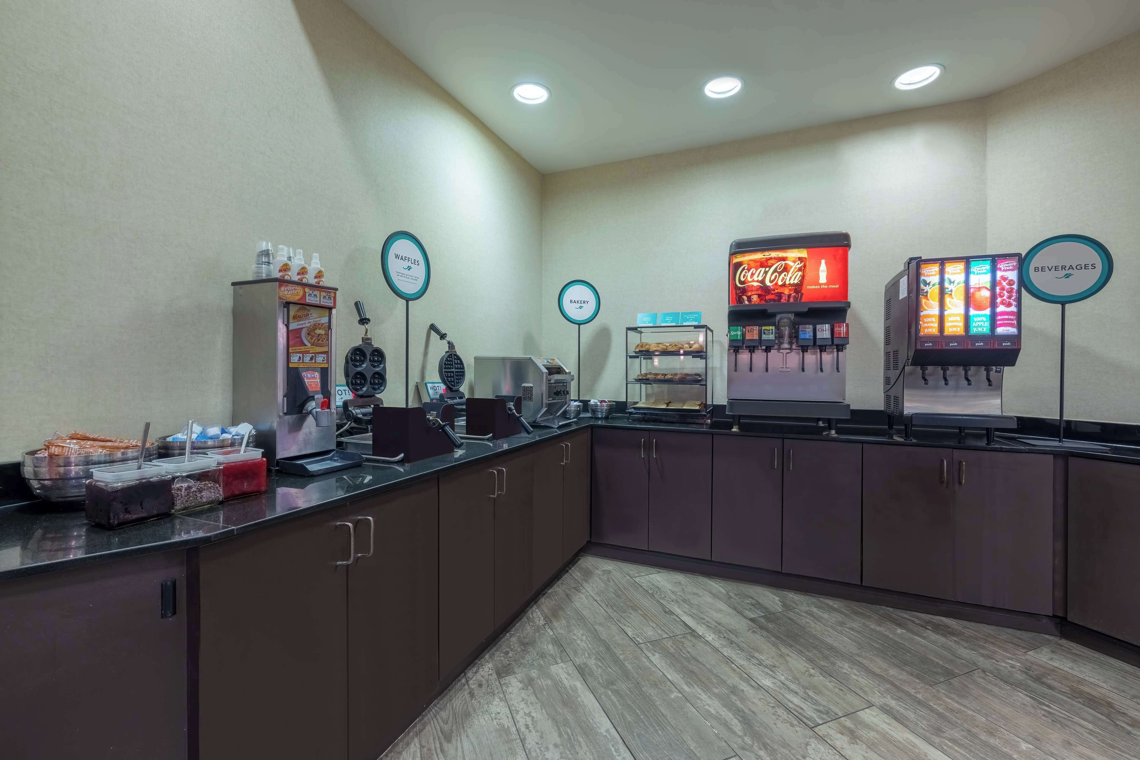 A self-service breakfast area with waffle makers, a Coca-Cola cooler, and a beverage dispenser.