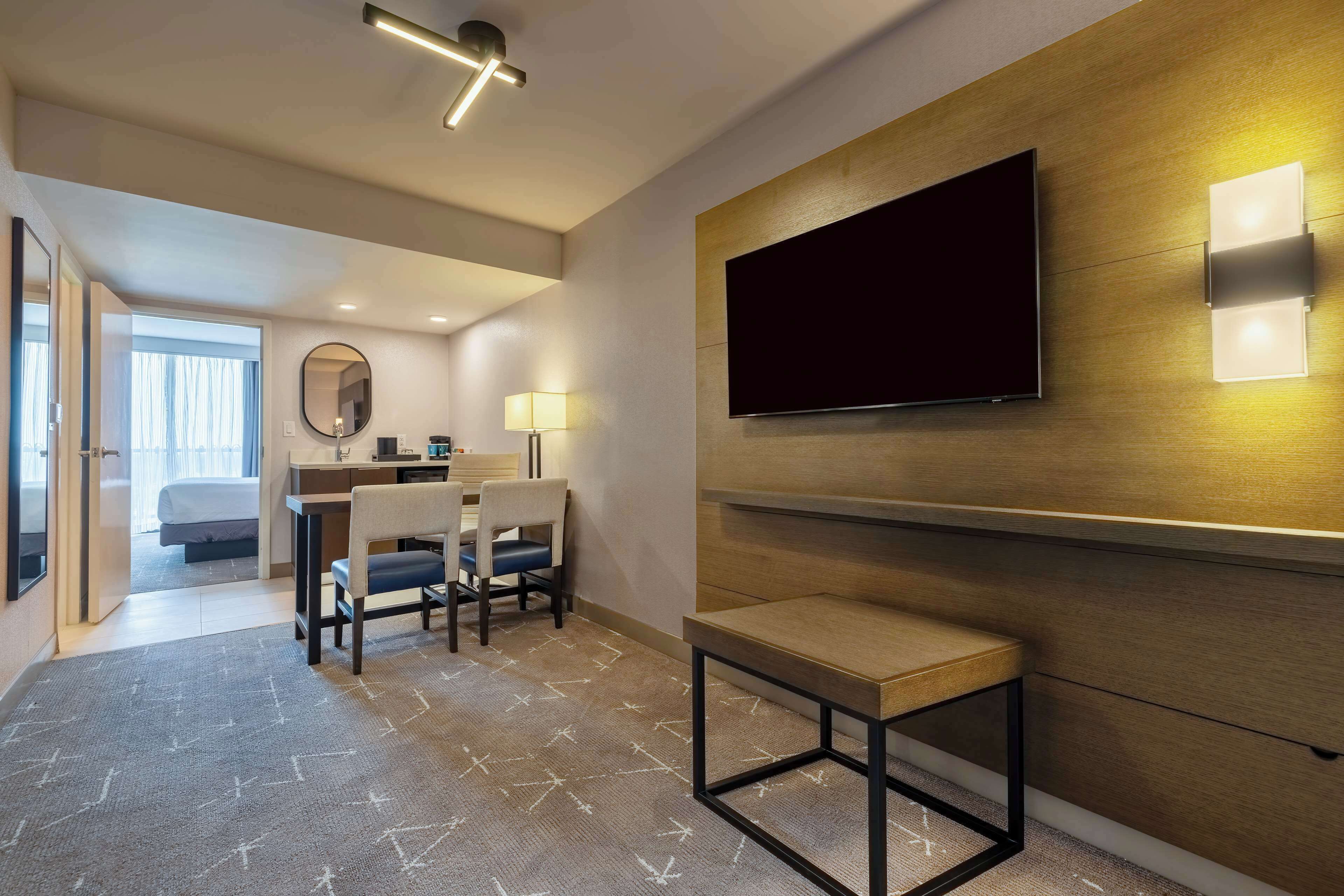 Een moderne hotelkamer met een grote flatscreen-televisie aan de muur, een bureau met stoel, een klein tafeltje en een bed op de achtergrond.