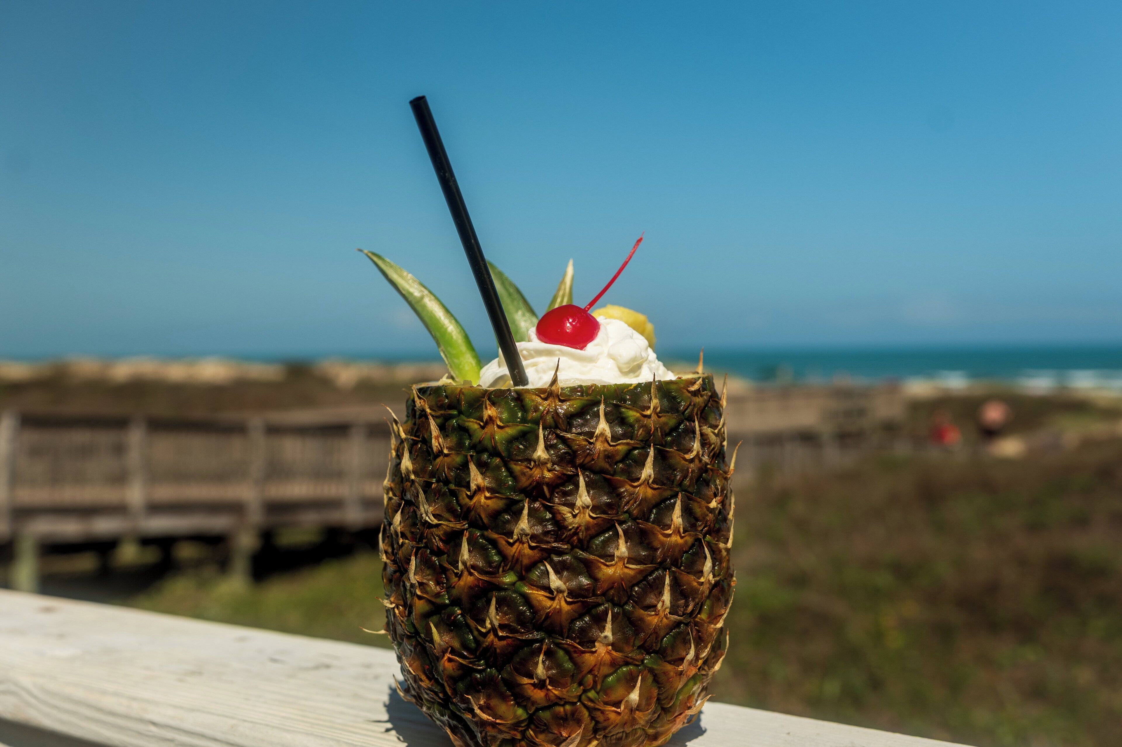 A pineapple drink with a cherry and straw on a wooden surface with a wooden fence and ocean in the background.