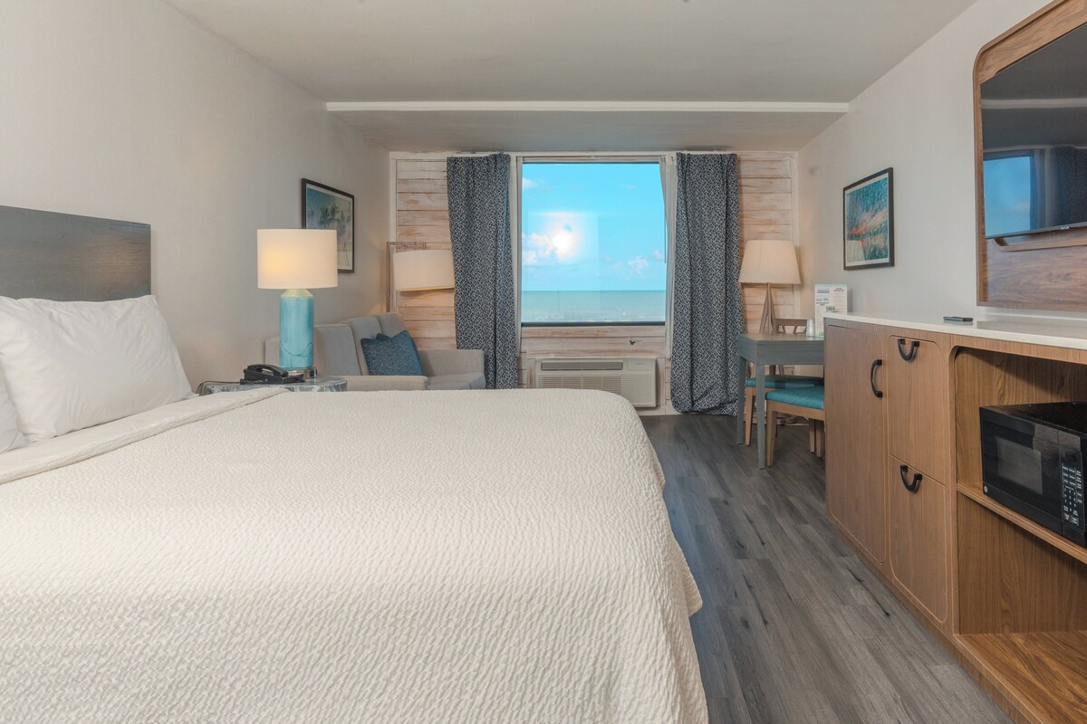 A hotel room with a large bed, a desk, a chair, a microwave, and a view of the ocean.
