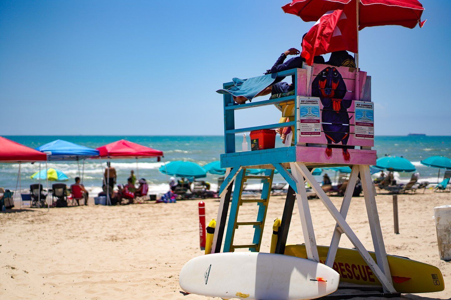 Una torretta di salvataggio con un ombrellone rosso e blu, una tavola da surf e un cartello che indica correnti pericolose.