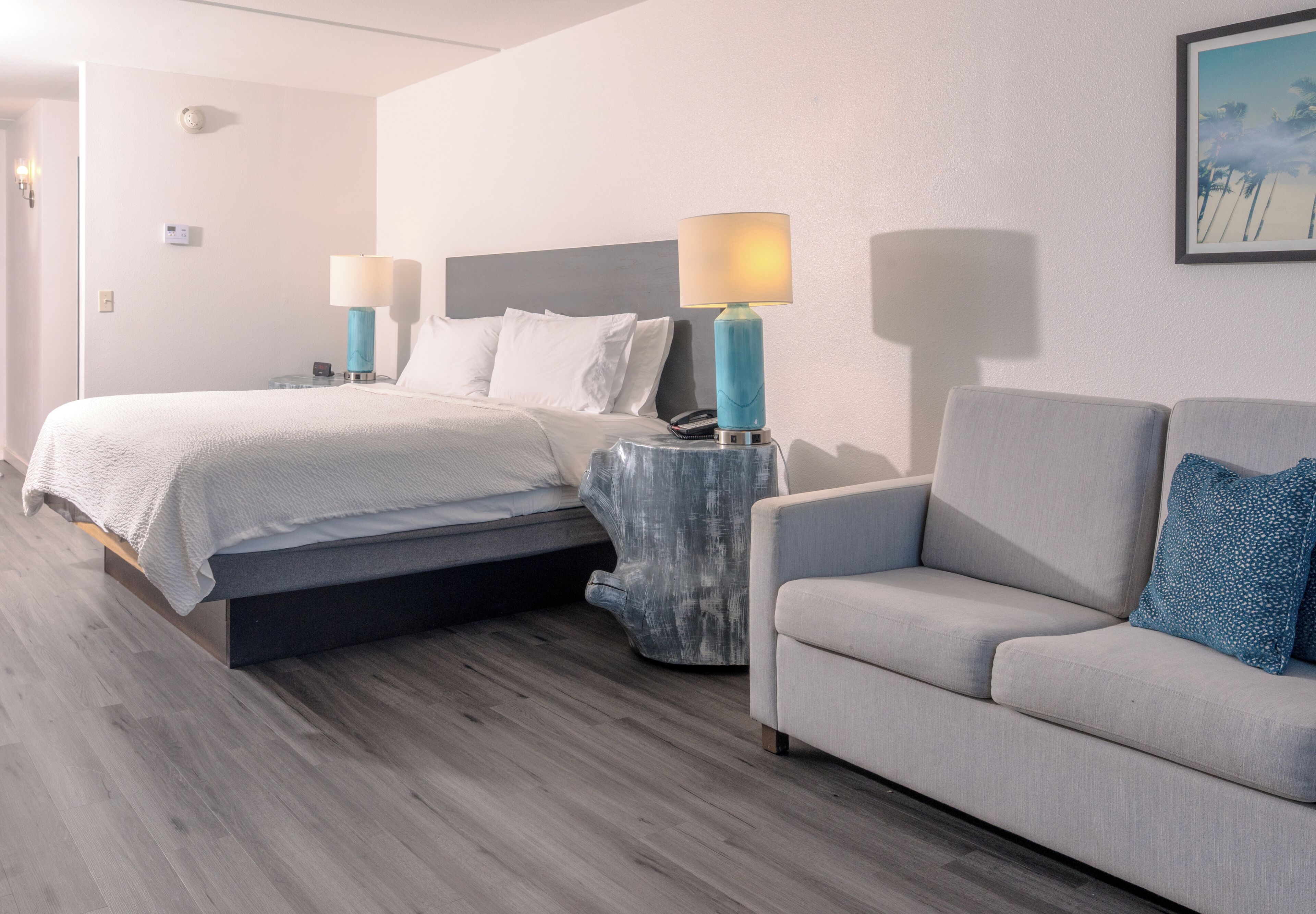 A hotel room with a bed, a sofa, two bedside tables with lamps, a wall-mounted clock, and a framed picture on the wall.