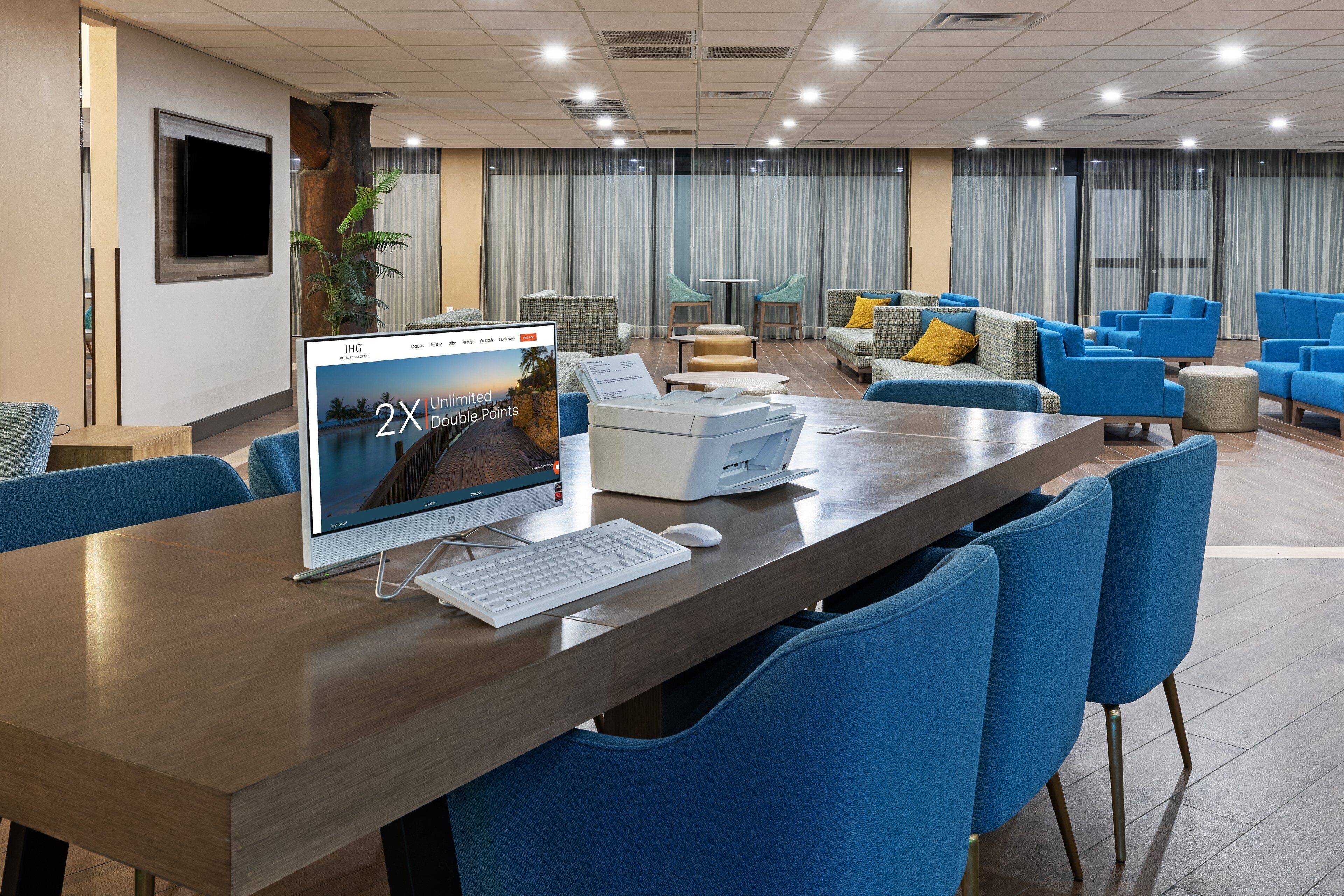 A modern hotel lobby with a wooden reception desk, a computer monitor, and blue chairs.