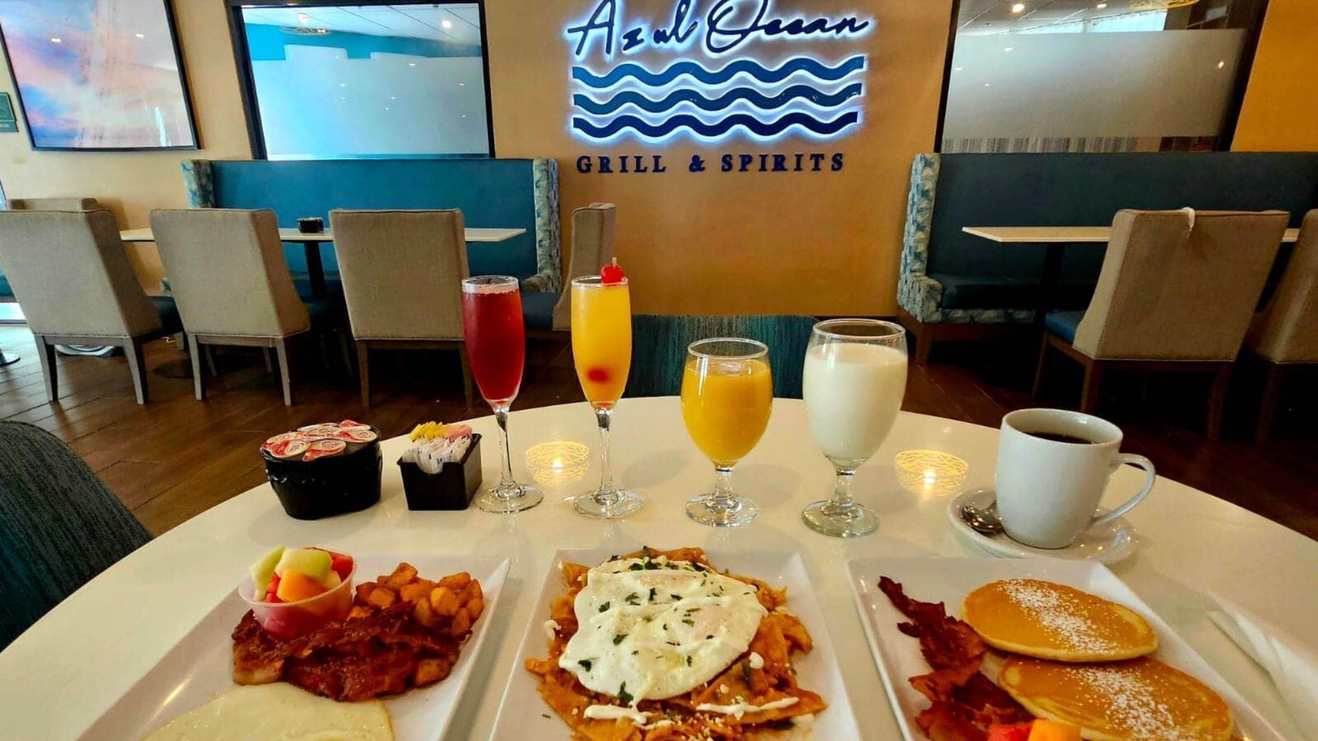 A breakfast setting with plates of food, drinks, and a neon sign reading "Azul Caribbean GRILL & SPIRITS".