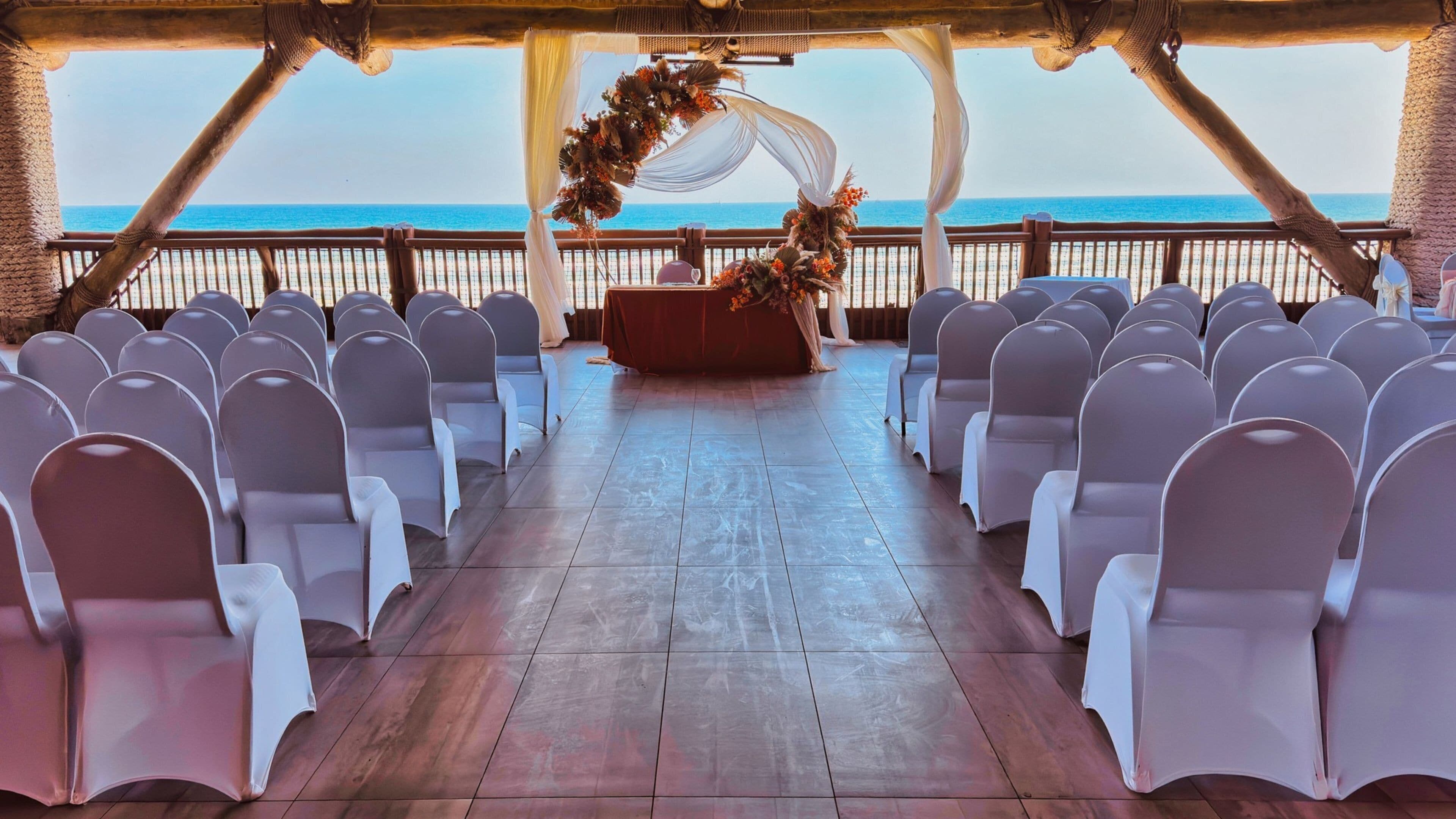 An aisle with rows of white chairs, a wooden floor, and a view of the ocean.