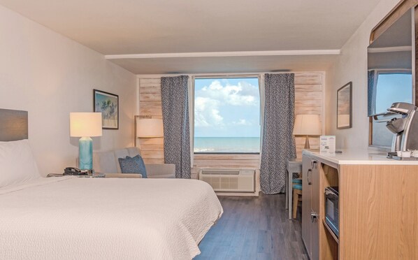 Habitación de hotel con cama, escritorio con computadora, vista al mar y ventana con cortinas.