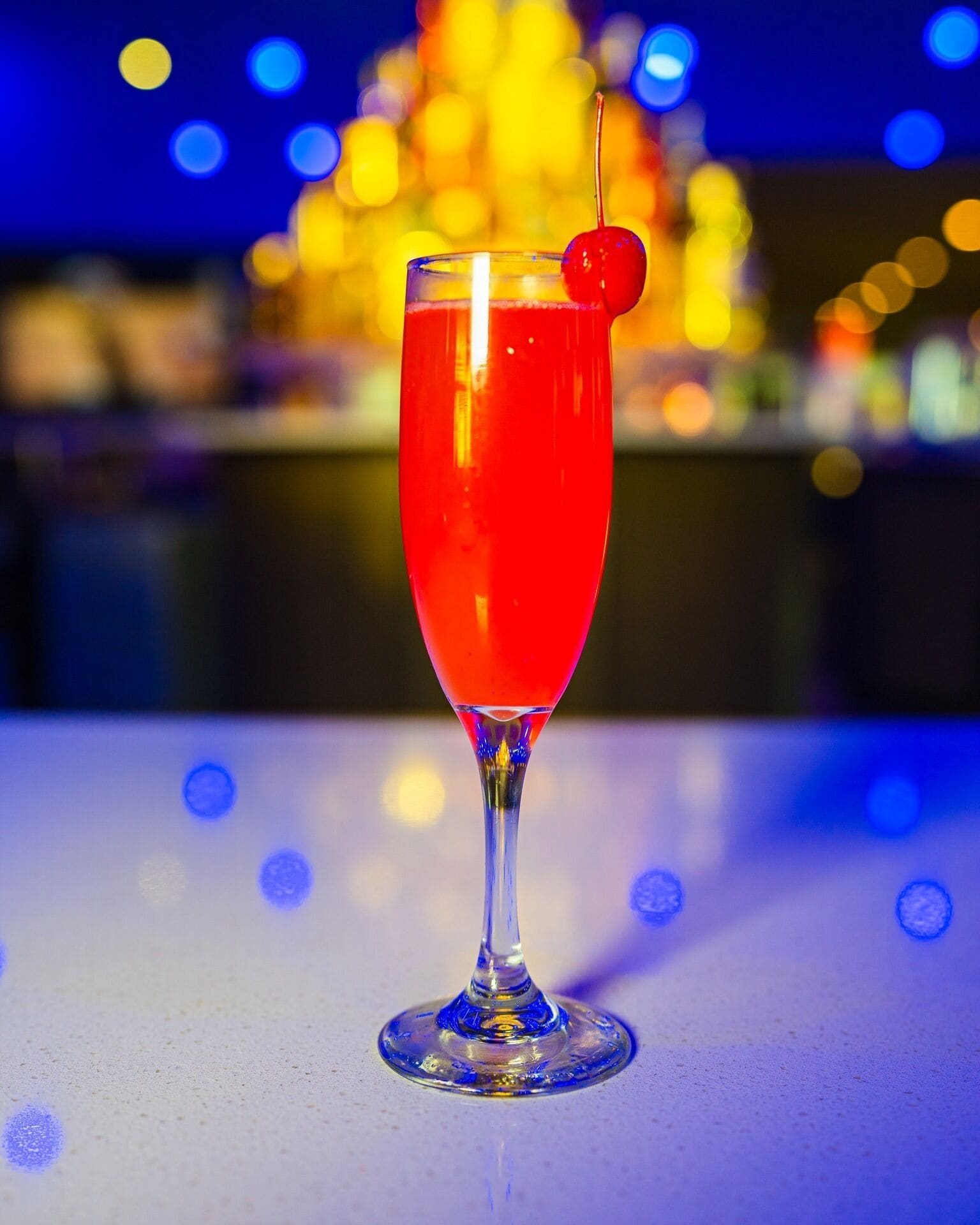 Et cocktailglas med et kirsebær på en pind, stående på en bardisk med et sløret baggrund af lys.