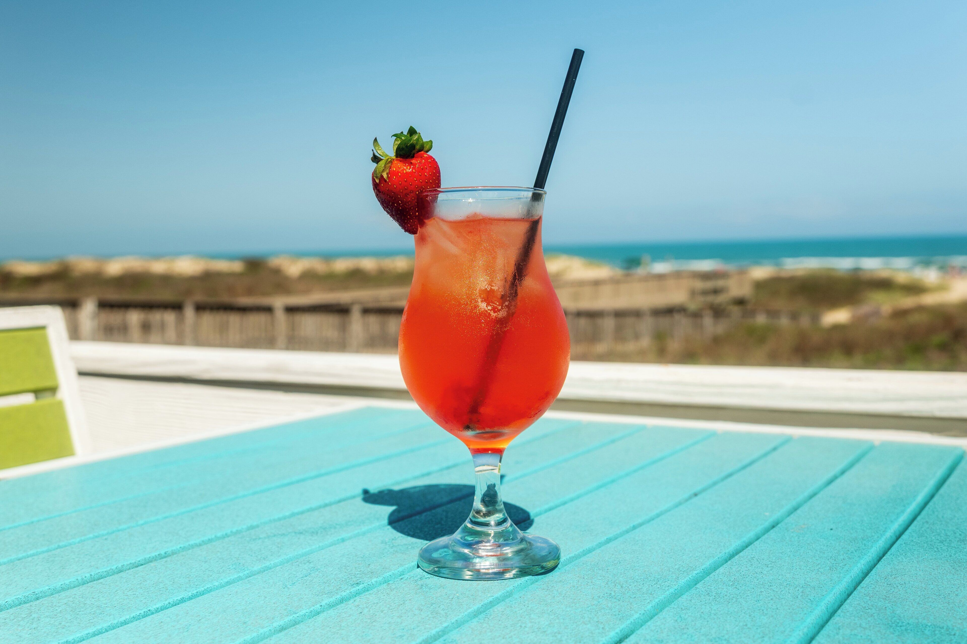 Un verre à cocktail, décoré d’une fraise, est posé sur une table bleue, avec une vue sur la plage en arrière-plan.