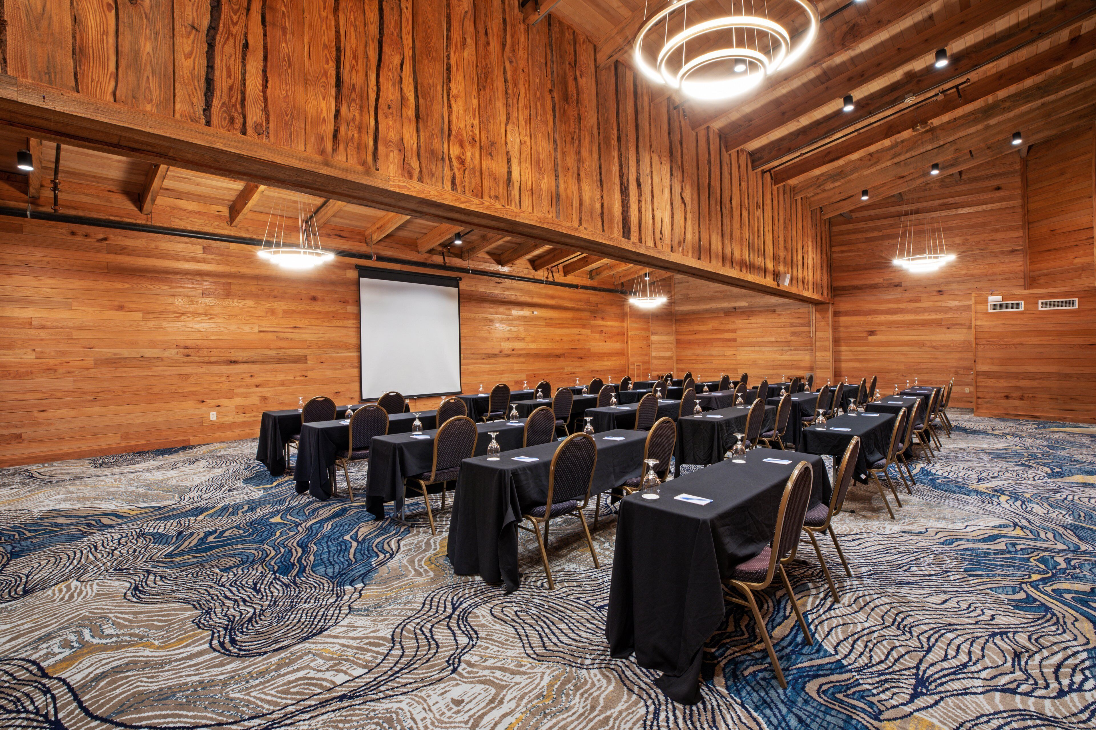 Uma sala de conferências com paredes de madeira, uma tela de projetor, mesas com cadeiras e um tapete estampado.