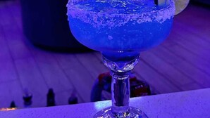 Zona de bar con un cóctel azul en un vaso, una rodaja de lima como decoración y una barra con diferentes botellas y un adorno decorativo.