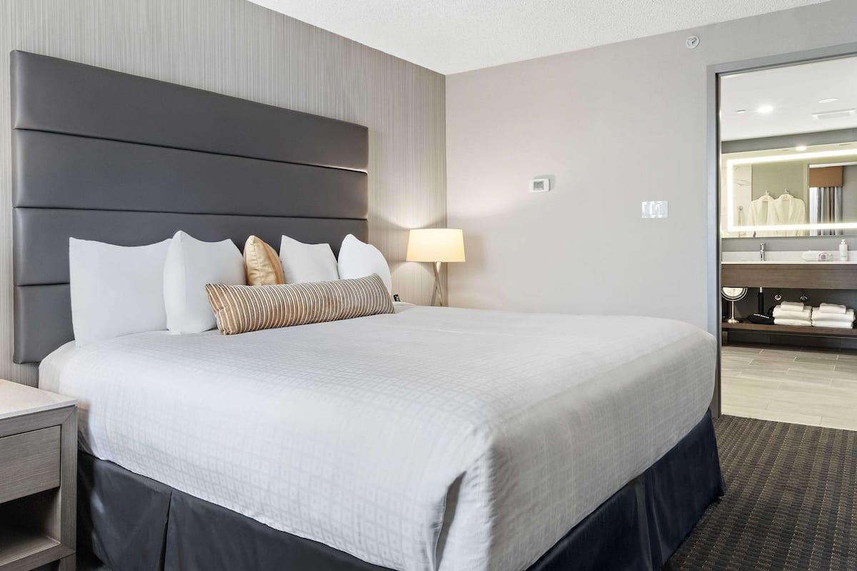 Une chambre d’hôtel avec un grand lit, une table de chevet, une lampe, un miroir et une salle de bain visible à travers une porte ouverte.