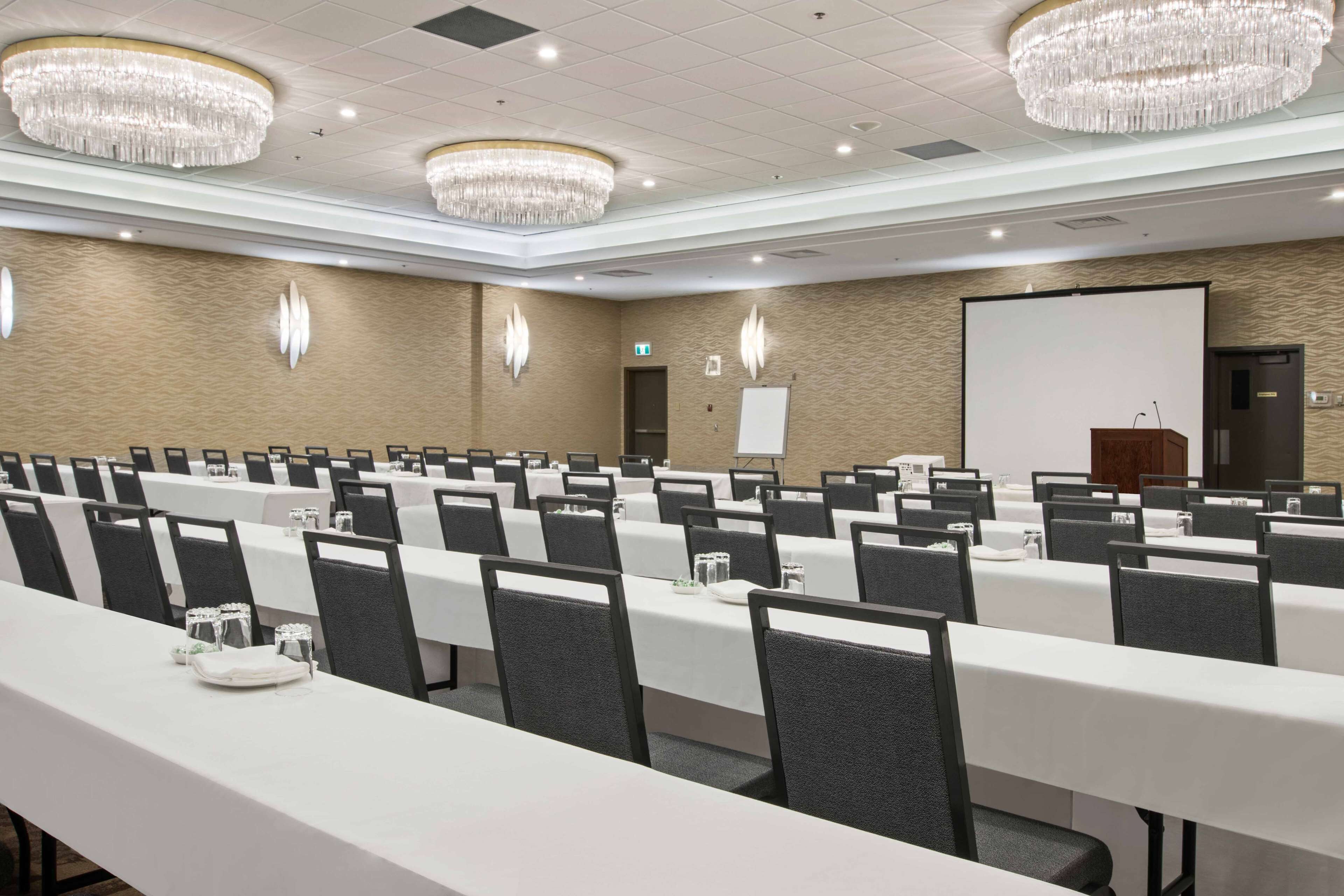 A conference room with long tables, chairs, chandeliers, a whiteboard, and a projector screen.