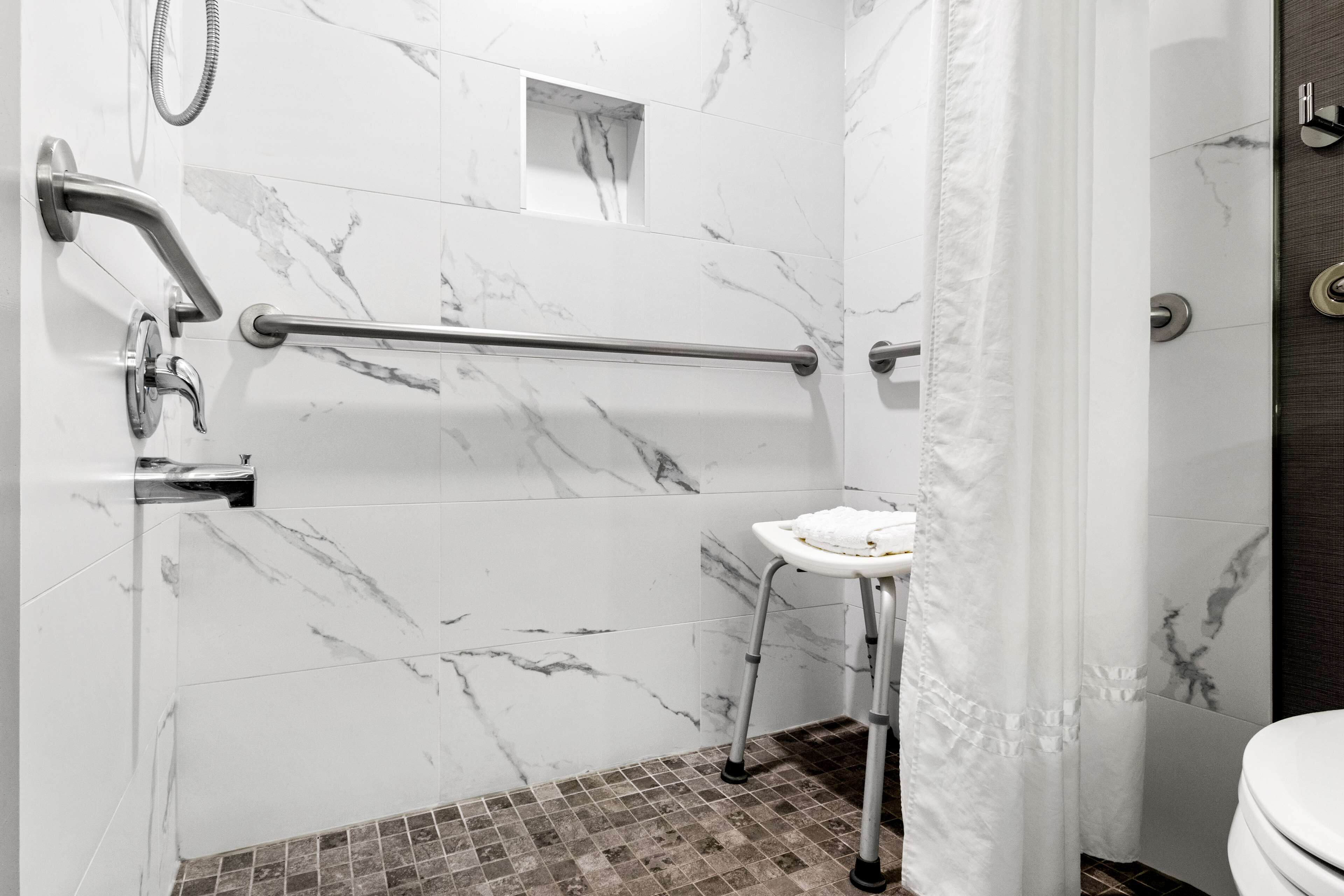 A bathroom with a white marble wall, a shower with a handheld showerhead, a grab bar, a small white sink, and a toilet.
