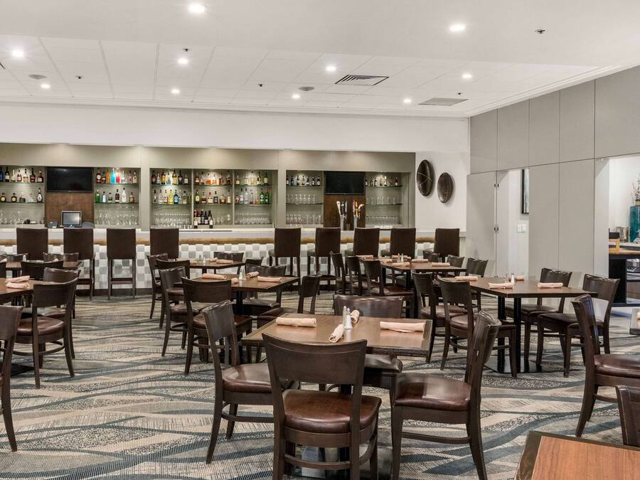 A hotel dining area with multiple tables and chairs, a bar with shelves of bottles, and a television screen.