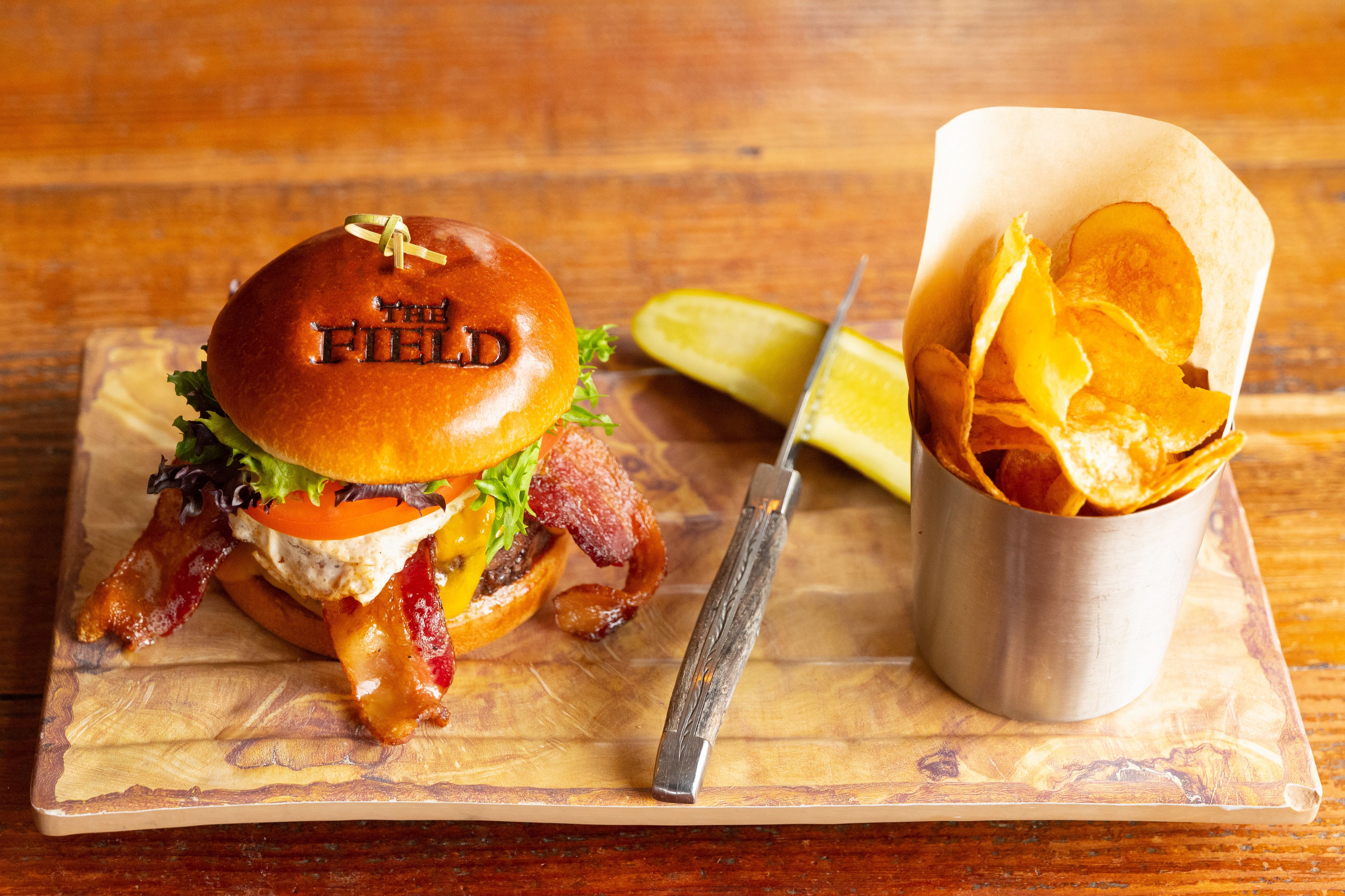 Ein Burger mit Speck, Salat, Tomate und einem Brötchen mit der Aufschrift 'The Field' auf einer Holzplatte.