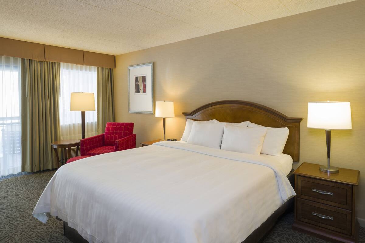 A hotel room with a large bed, two bedside tables with lamps, a red armchair, and a window with curtains.