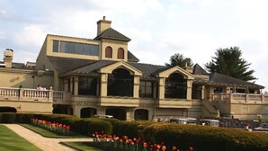 A large, multi-story building with a prominent entrance, a covered patio area, and a well-maintained garden with red flowers.