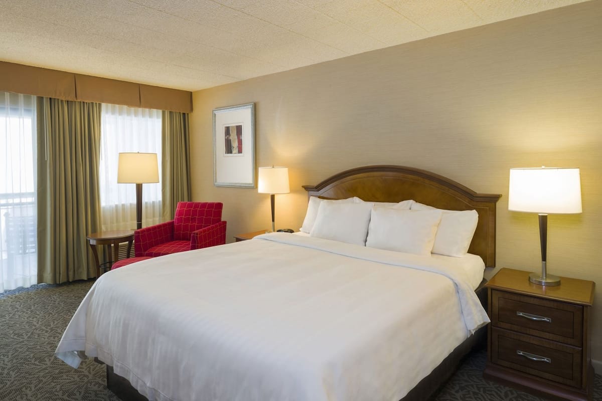 A hotel room with a large bed, two bedside lamps, a red armchair, a small table, and a framed picture on the wall.