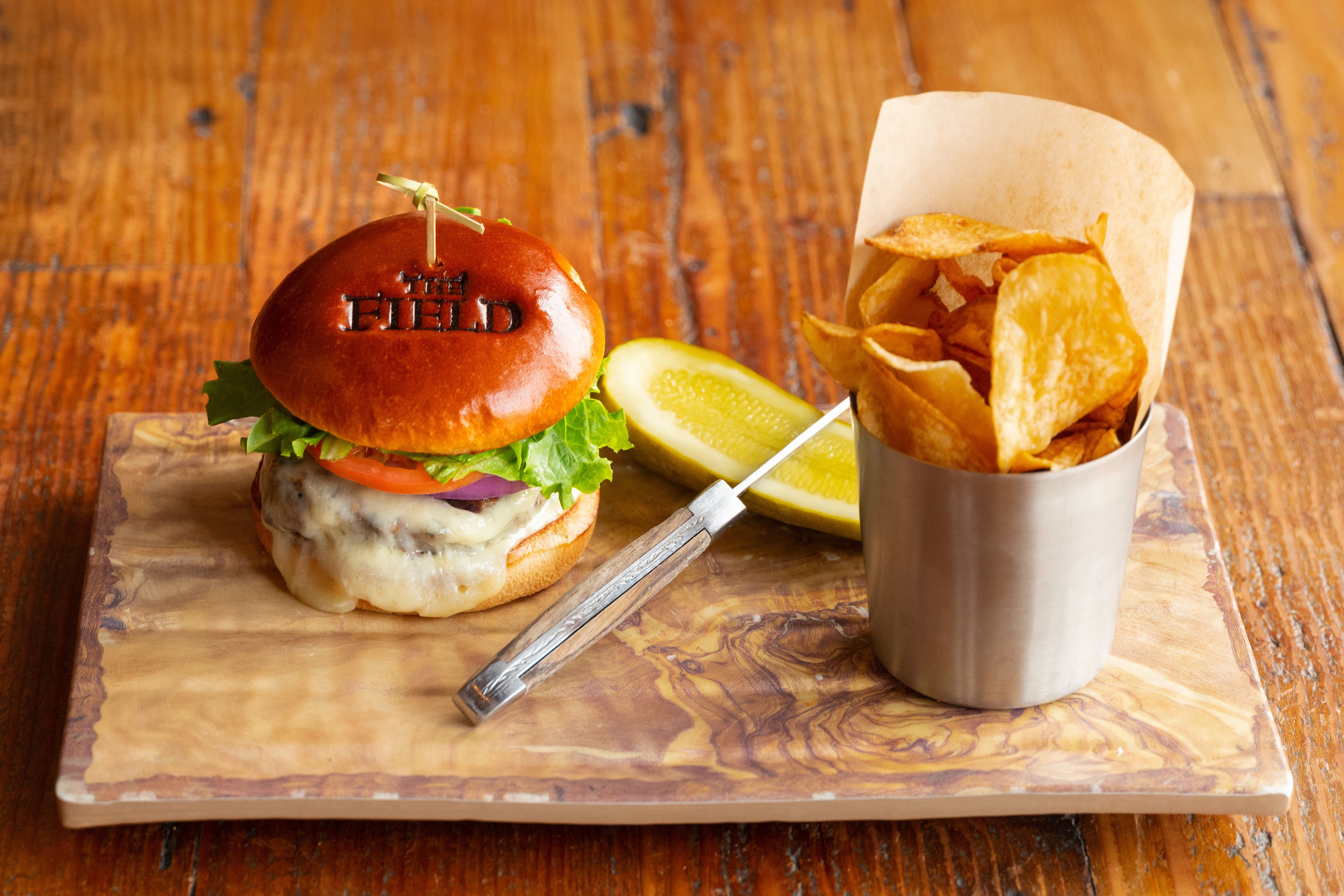 Un hamburger servi dans un pain marqué du nom « THE FIELD », présenté sur une planche en bois, accompagné d’une portion de frites et d’une tranche de citron.