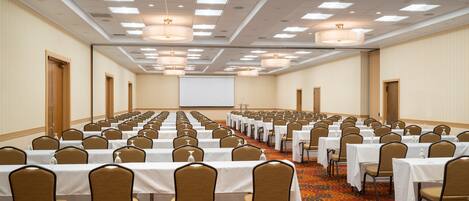 A conference room with rows of tables and chairs, a large screen at the front, and a carpeted floor.
