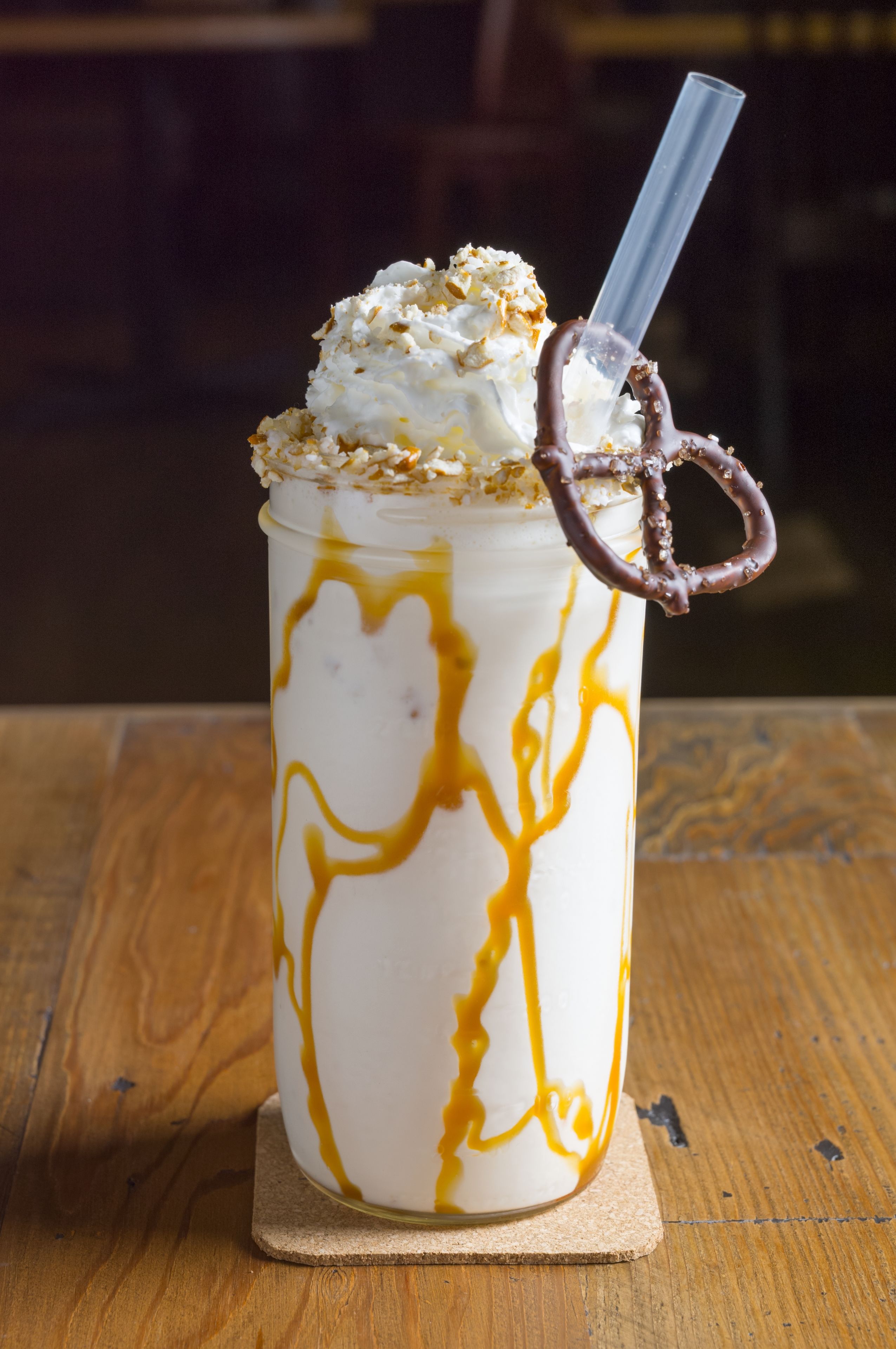 Une boisson dessert servie dans un grand verre avec une paille, surmontée de crème fouettée et d’un anneau de chocolat, le tout posé sur un sous-verre en bois.