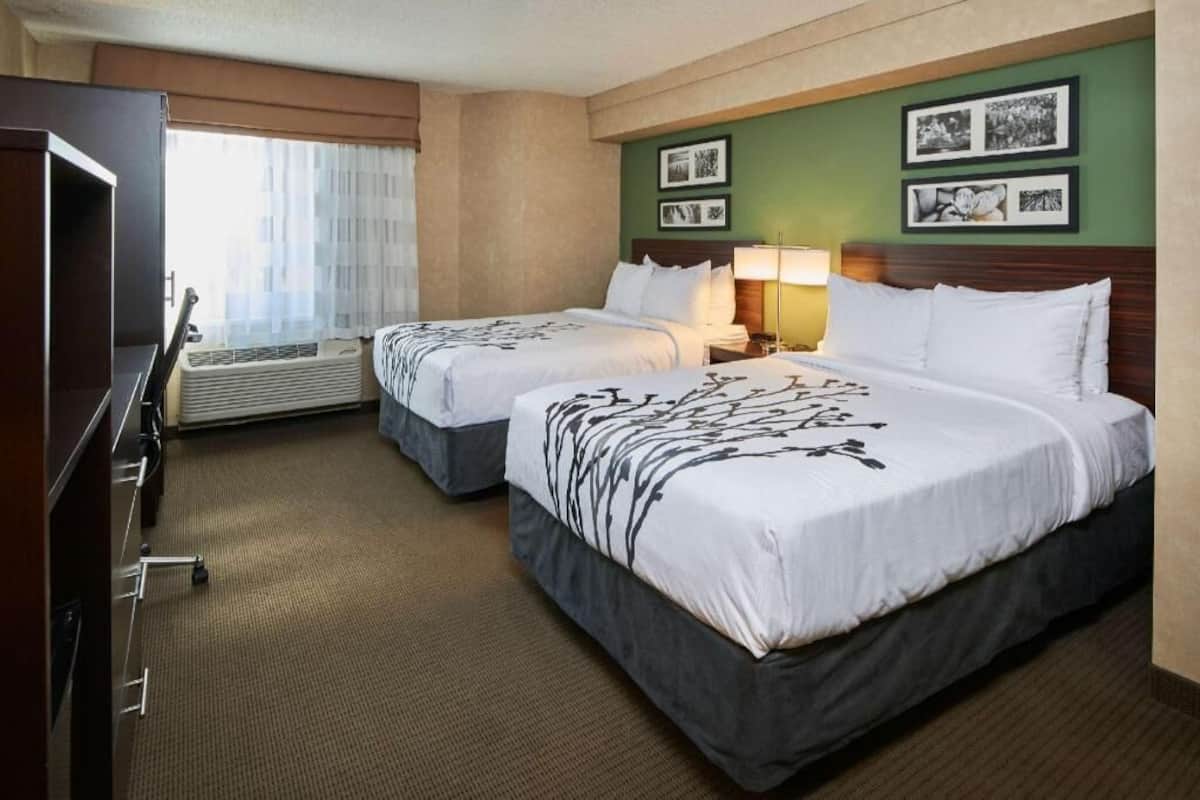 A hotel room with two beds, a desk, an air conditioning unit, and framed pictures on the wall.
