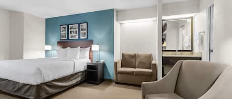 Una habitación de hotel con cama, mesita de noche, lámpara, un sofá, una silla y un baño visible a través de una puerta abierta.