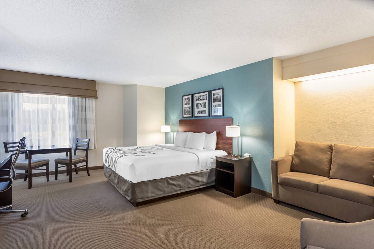 A hotel room with a bed, bedside tables, a desk with chairs, a couch, and framed pictures on the wall.