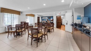 A hotel dining area with multiple tables and chairs, a kitchen in the background, and an exit sign.