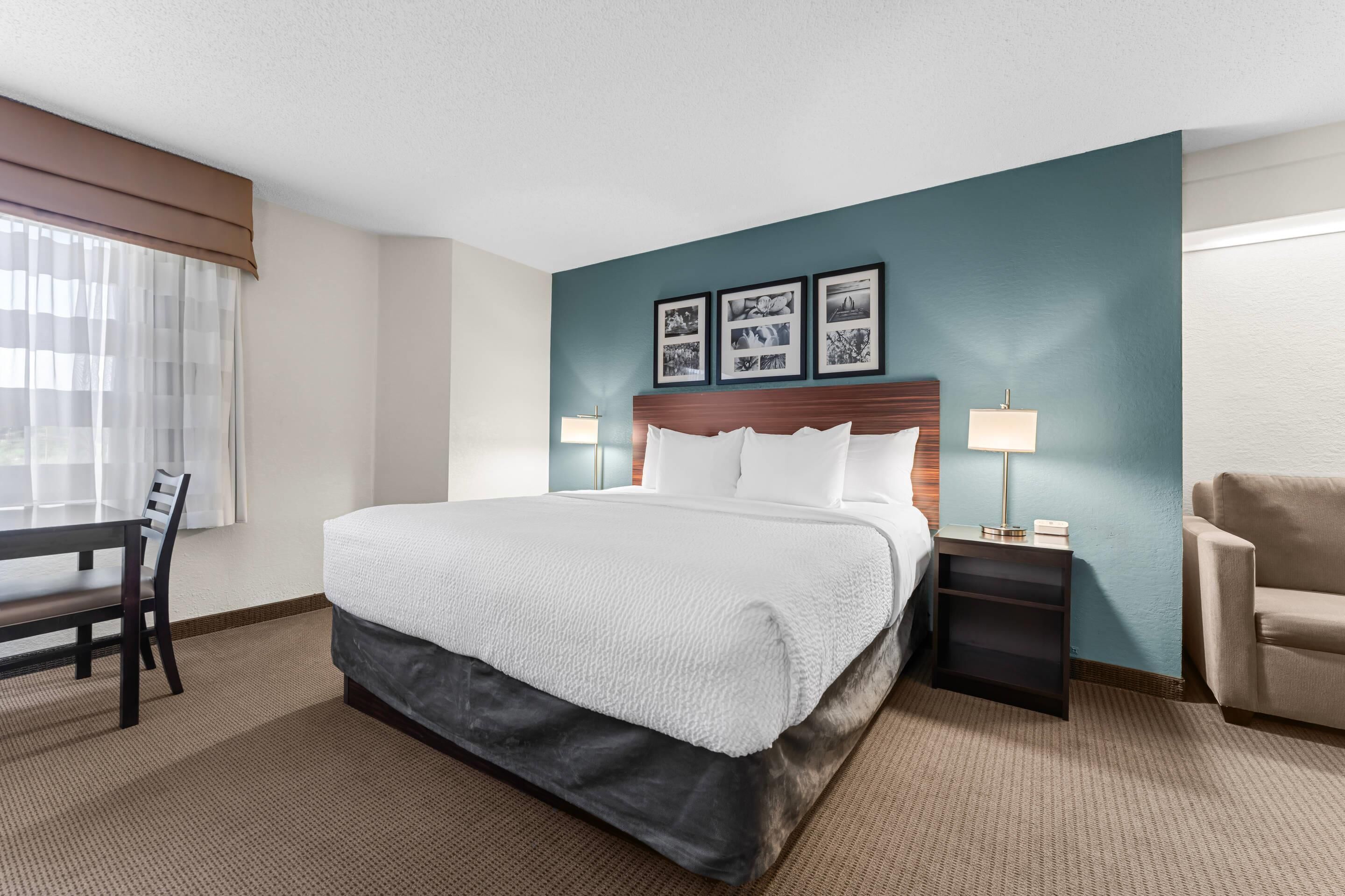 A hotel room with a bed, a desk, a chair, a nightstand, and a lamp.