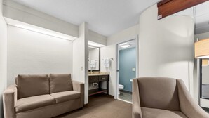 A hotel room with a sofa, a chair, a bathroom with a toilet and sink, and a mirror.