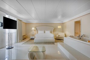 A modern hotel room with a large bed, a sitting area, a bathtub, and a flat-screen TV.
