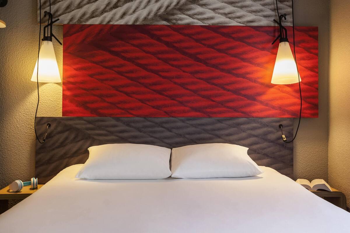 Habitación de hotel con cama, dos lámparas de noche, mesita con teléfono y pared con diseño geométrico.