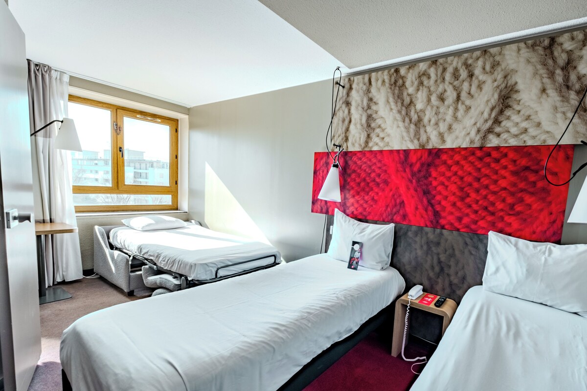 Habitación de hotel con cama, mesita de noche con teléfono, ventana con cortinas y cabecero con diseño rojo y beige.
