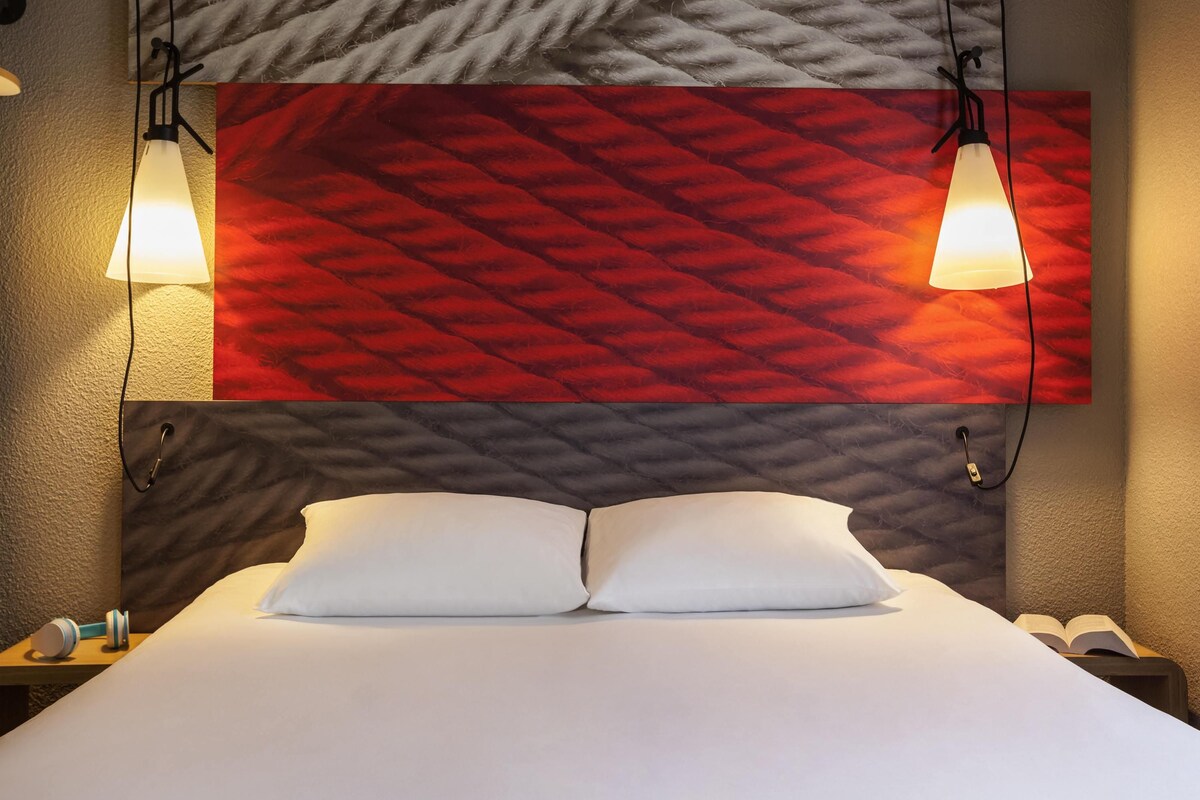 A hotel room with a bed, two bedside lamps, a nightstand with a phone, and a wall with a geometric patterned design.