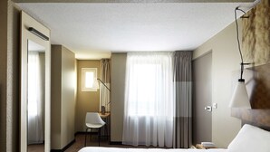 Una habitación de hotel con una cama grande, una silla, un escritorio, una lámpara y un ventanal con cortinas.