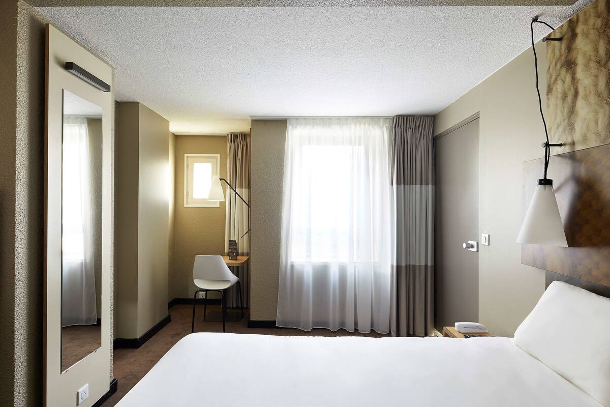 A hotel room with a large bed, a chair, a desk, a lamp, and a window with curtains.