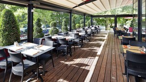 An outdoor patio with tables and chairs, a covered area, and a view of the street and greenery.