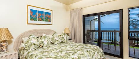 A bedroom with a bed, a nightstand, a lamp, a framed picture, and a sliding glass door leading to a balcony with a view of the ocean.