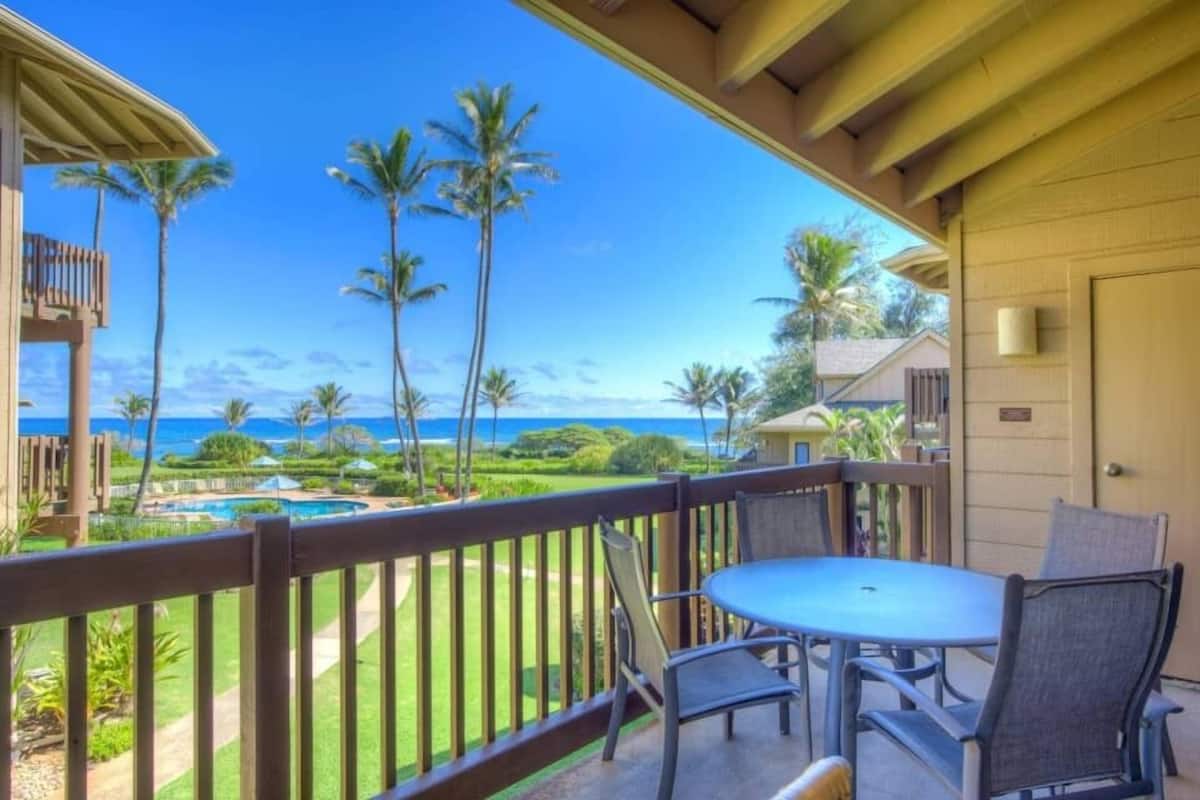 A balcony with a view of the ocean, palm trees, and a pool.