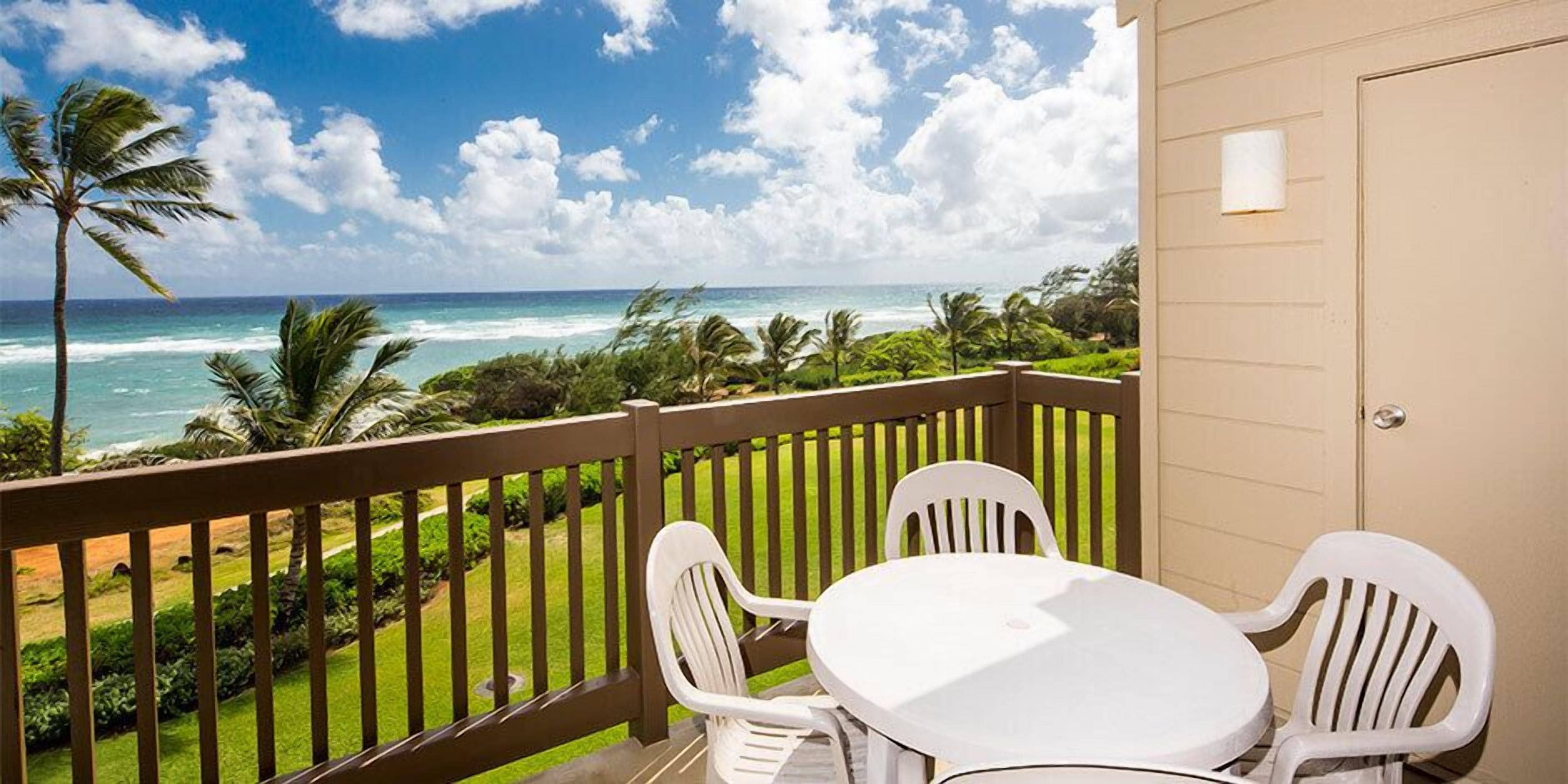 A balcony with a white table and chairs, overlooking a beach with palm trees and the ocean.