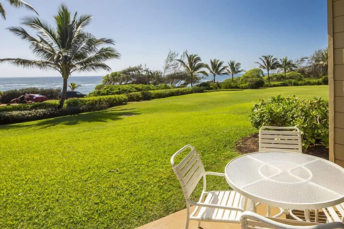 A patio with a round table and two chairs, overlooking a well-maintained lawn and palm trees, with a view of the ocean.