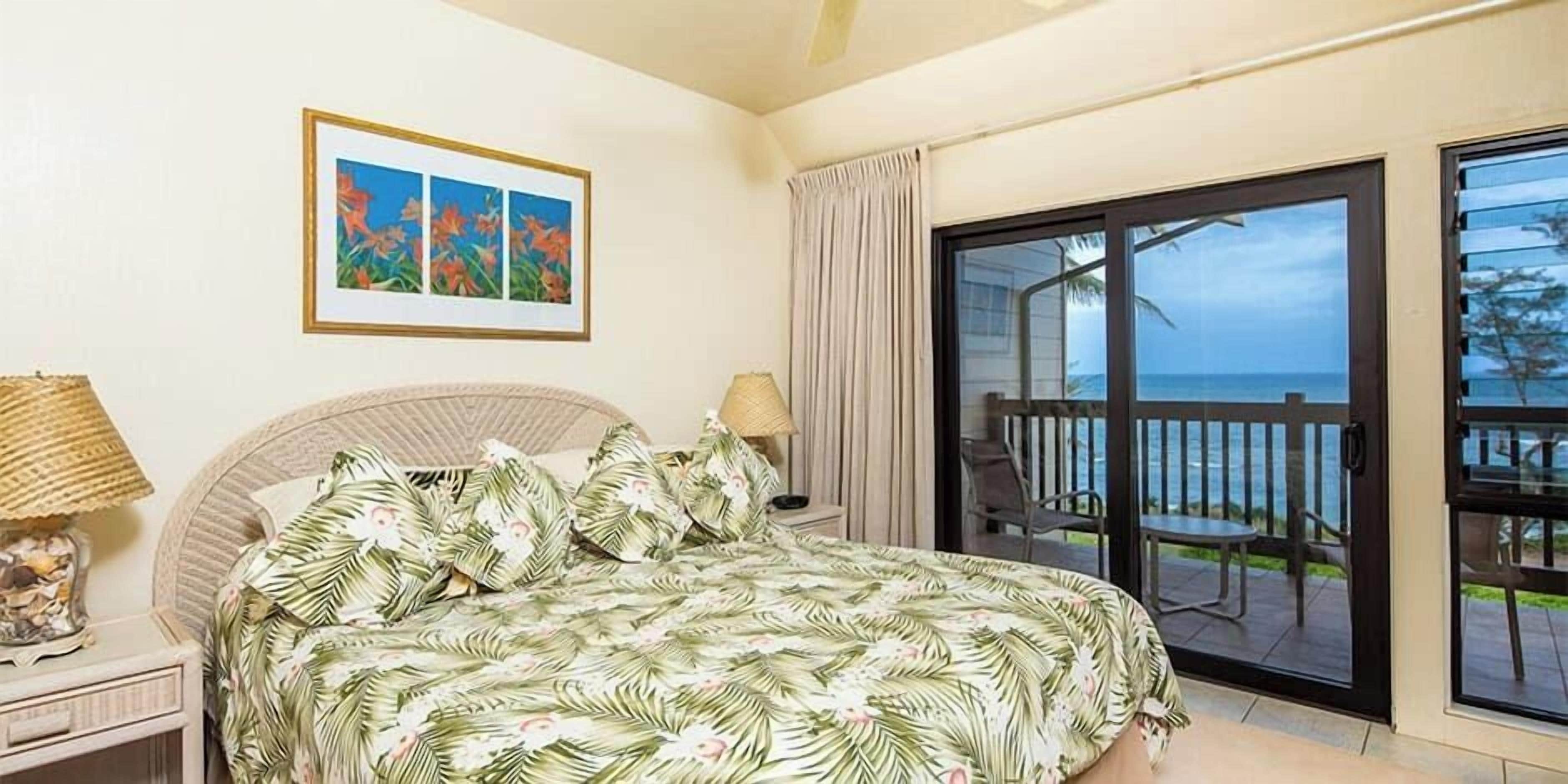 A bedroom with a bed, a nightstand, a lamp, a framed picture, and a balcony with a view of the ocean.