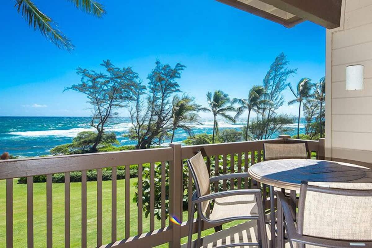A balcony with a round table and chairs, overlooking a beach with palm trees and the ocean.