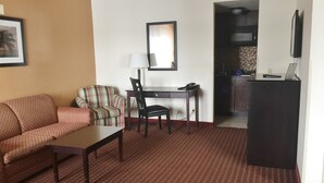 A hotel room with a sofa, a striped armchair, a desk with a lamp, and a small kitchenette.