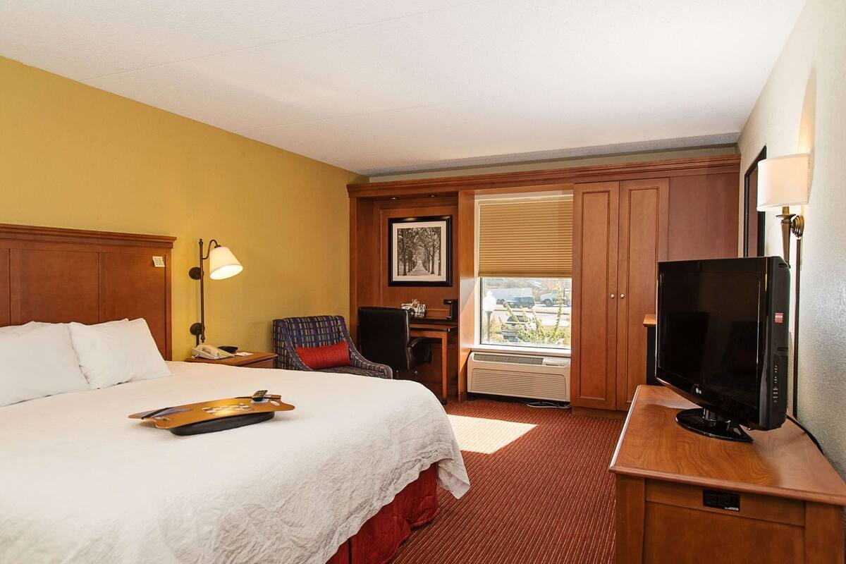 Ein Hotelzimmer mit Bett, Schreibtisch mit Fernseher, Stuhl, Lampe und einem Fenster mit Jalousien.