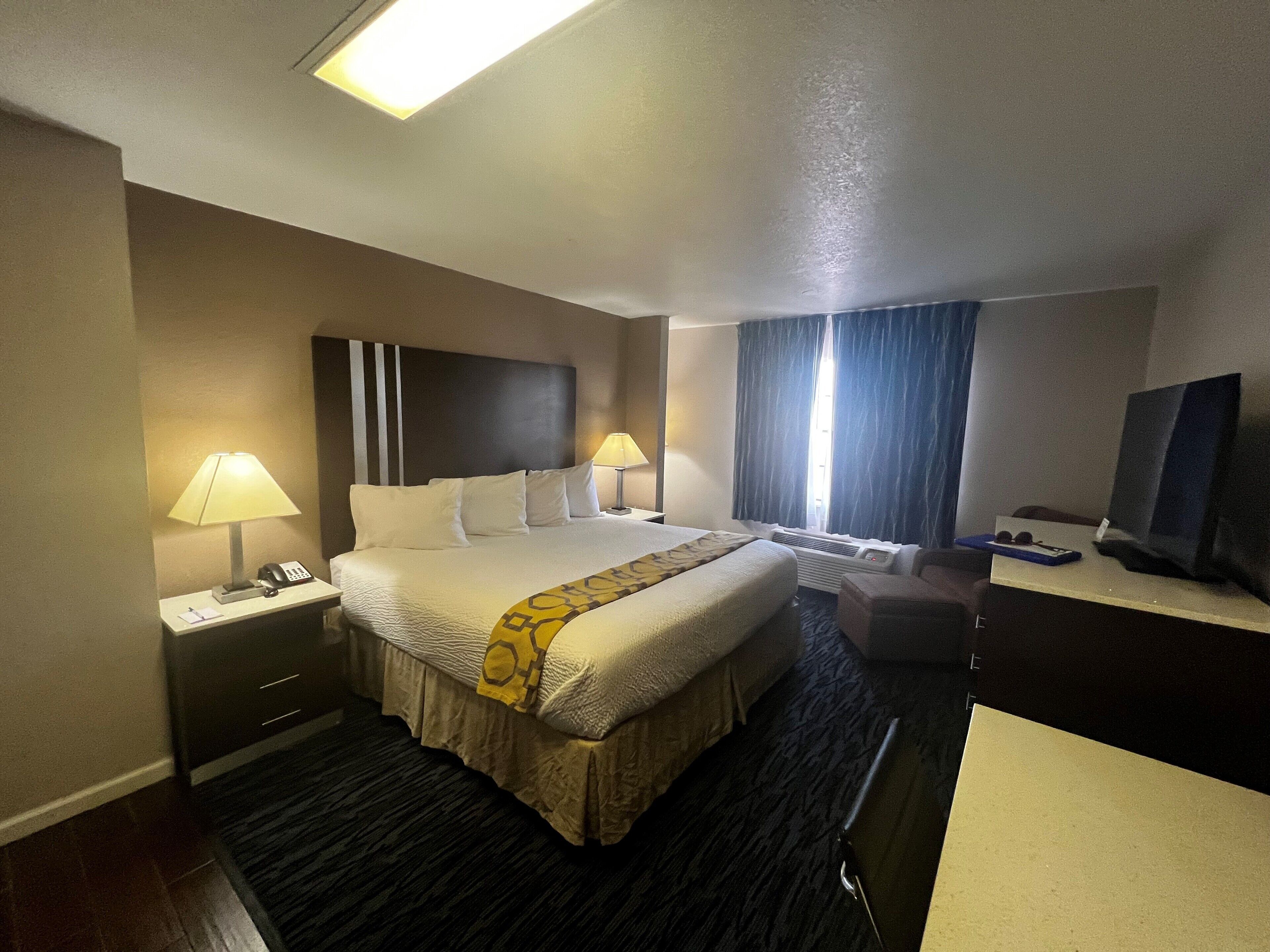 Quarto de hotel com uma cama grande, duas luminárias de cabeceira, uma escrivaninha com televisão e uma janela com cortinas.