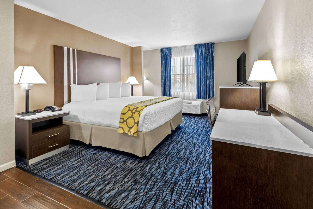 A hotel room with a large bed, two bedside tables with lamps, a TV on a stand, and a window with blue curtains.
