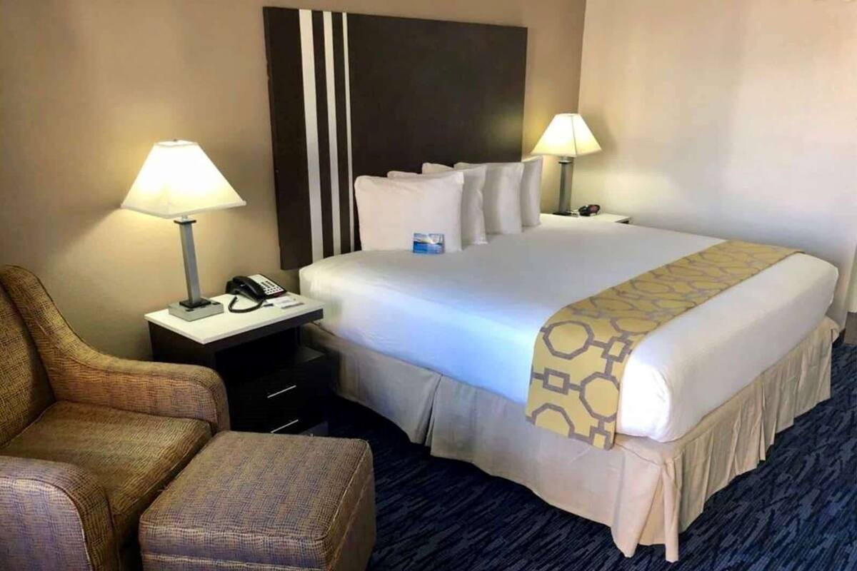 A hotel room with a bed, two bedside lamps, a chair, a small table with a phone, and a carpeted floor.