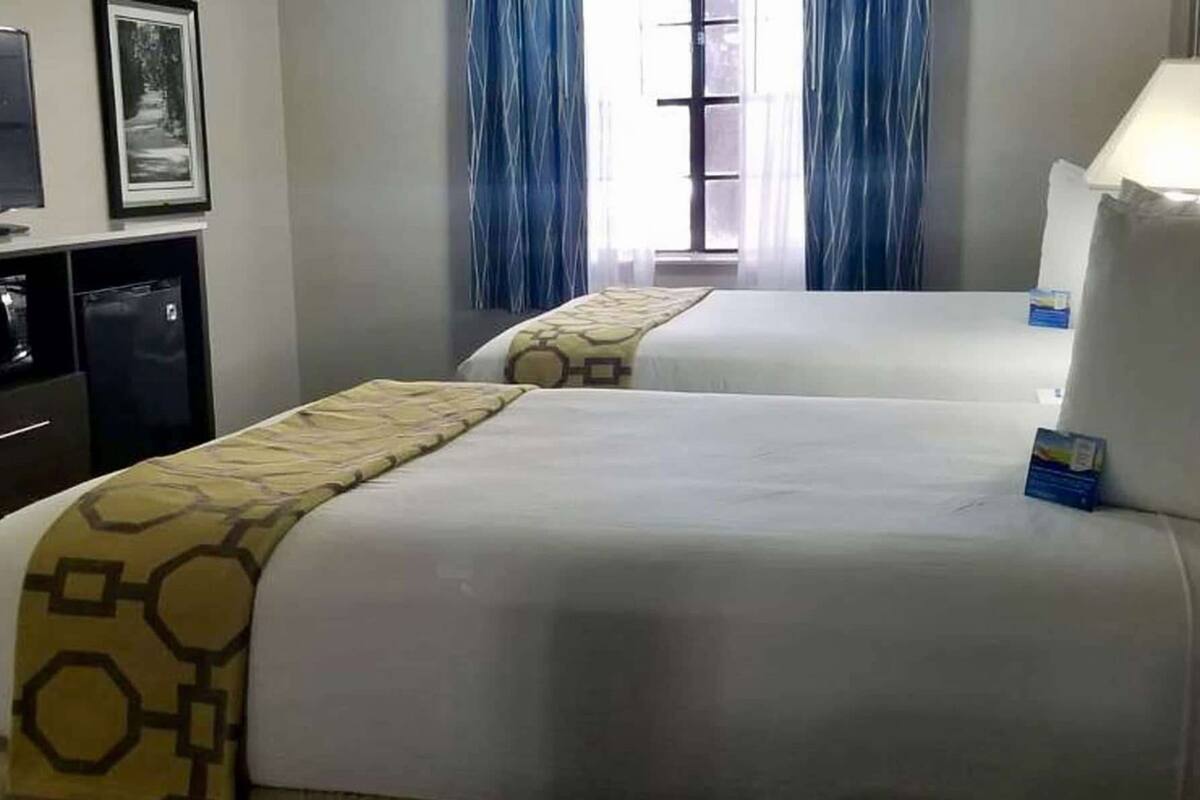 A hotel room with two beds, a desk, a small refrigerator, and a window with curtains.